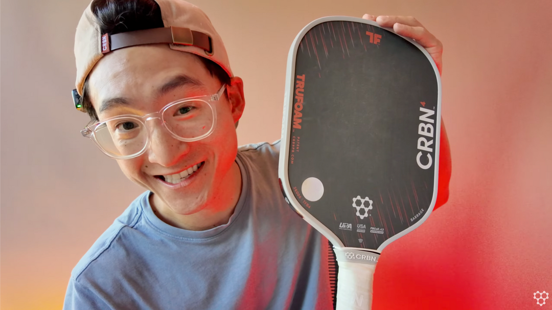 Smiling person in a baseball cap and clear glasses holds up a CRBN TruFoam Barrage pickleball paddle.
