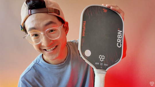 Smiling person in a baseball cap and clear glasses holds up a CRBN TruFoam Barrage pickleball paddle.